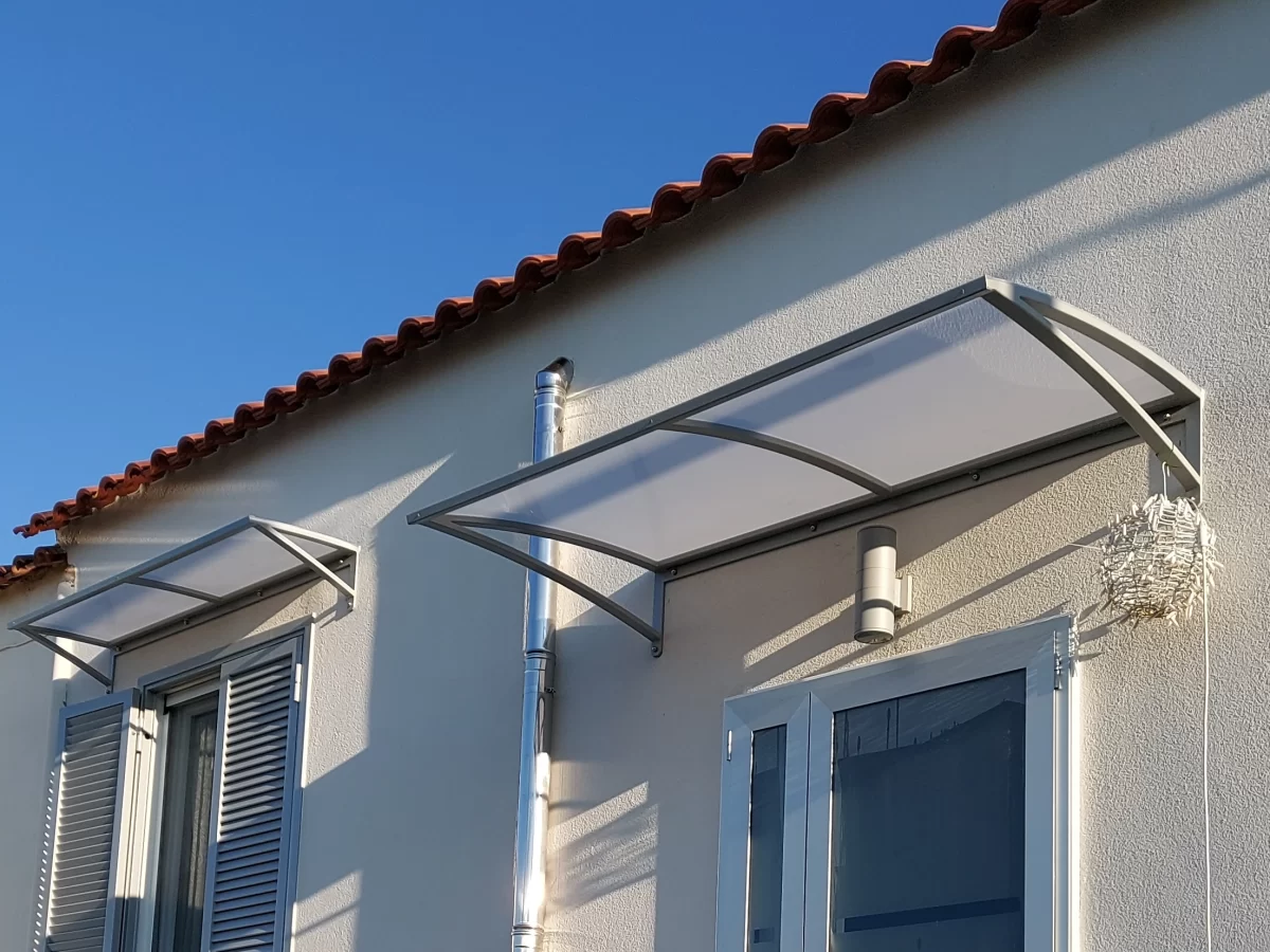 Pensilina curva con copertura in materiale sintetico traslucido e struttura portante in metallo, installata sopra una serie di finestre.
